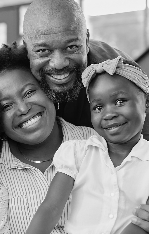 Smiling family portrait of two adults and a child. The child wears a headband bow. All are looking at the camera. Black and white image.