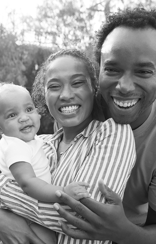A smiling couple holds their happy baby outdoors in a black-and-white photo.