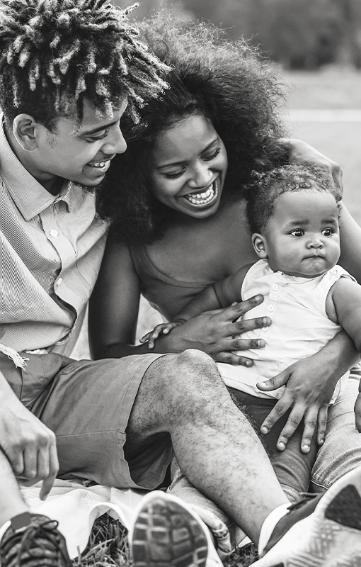 A family of three sits outdoors on a blanket, smiling and interacting with their baby.