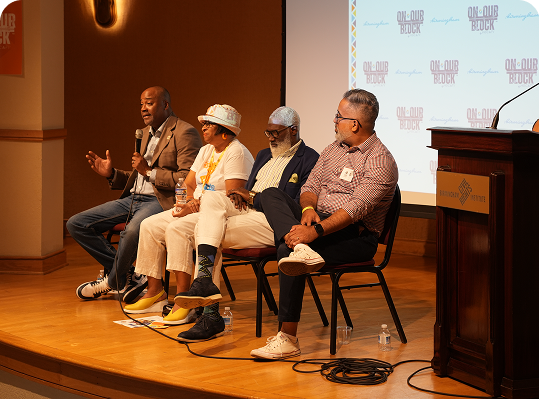 Four people sit on a stage panel, speaking into microphones during an event. A projected screen and event signage are visible in the background. Four people sit on a stage panel, speaking into microphones during an event. A projected screen and event signage are visible in the background.