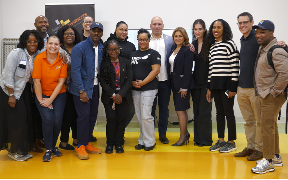 A group of fifteen adults pose together indoors for a group photo, standing on a yellow and green floor in front of a white wall and banner. A group of fifteen adults pose together indoors for a group photo, standing on a yellow and green floor in front of a white wall and banner.