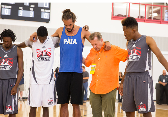 Five people stand on a basketball court with arms around each other, heads bowed, appearing to share a moment of silence or reflection. Five people stand on a basketball court with arms around each other, heads bowed, appearing to share a moment of silence or reflection.