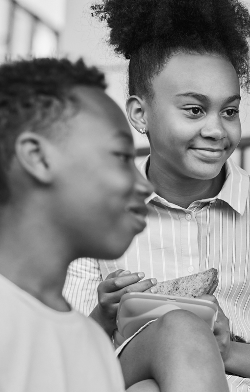 Black and white photo of a girl holding a sandwich and smiling, with a boy in the foreground, both sitting together indoors.