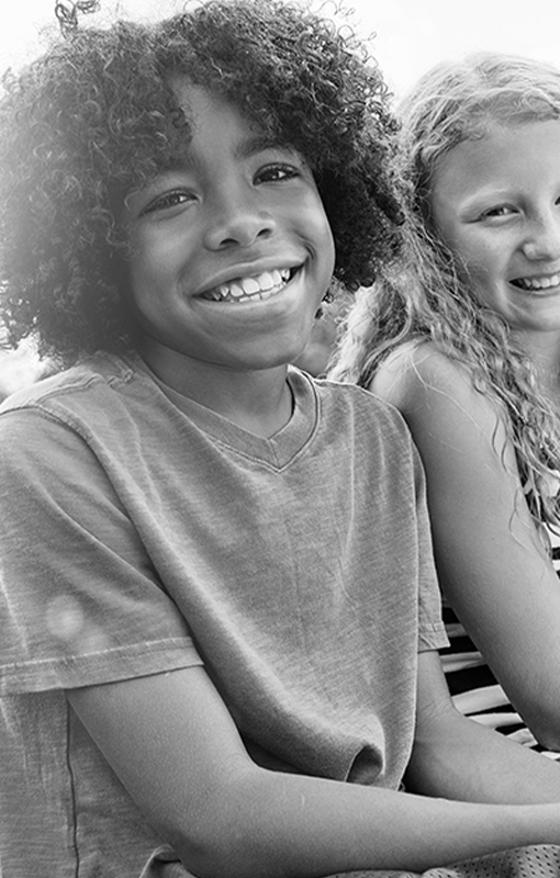 Two children sitting side by side outdoors, smiling at the camera. The image is in black and white.