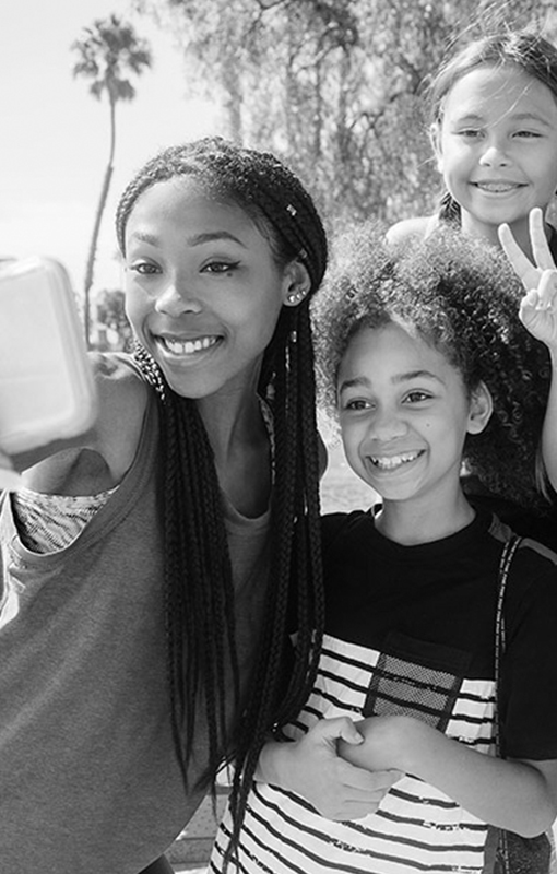 Three children pose for a selfie outdoors on a sunny day. One holds the phone, while another makes a peace sign. Trees and a palm tree are visible in the background.
