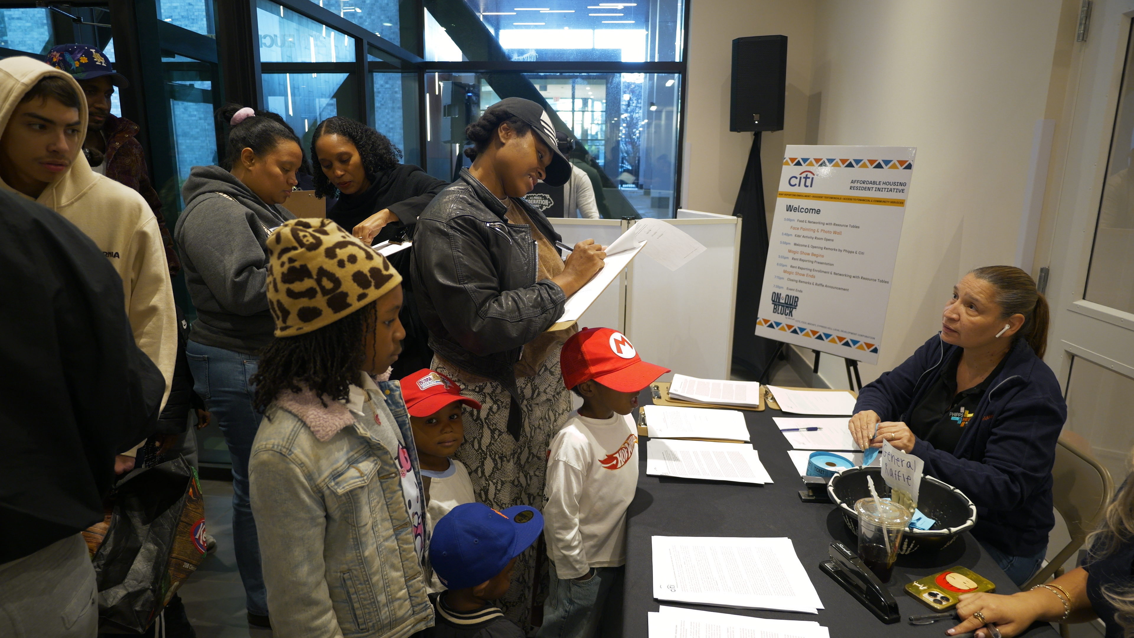 Children and adults play basketball together under a tent outside a building, while a man stands near the entrance watching.