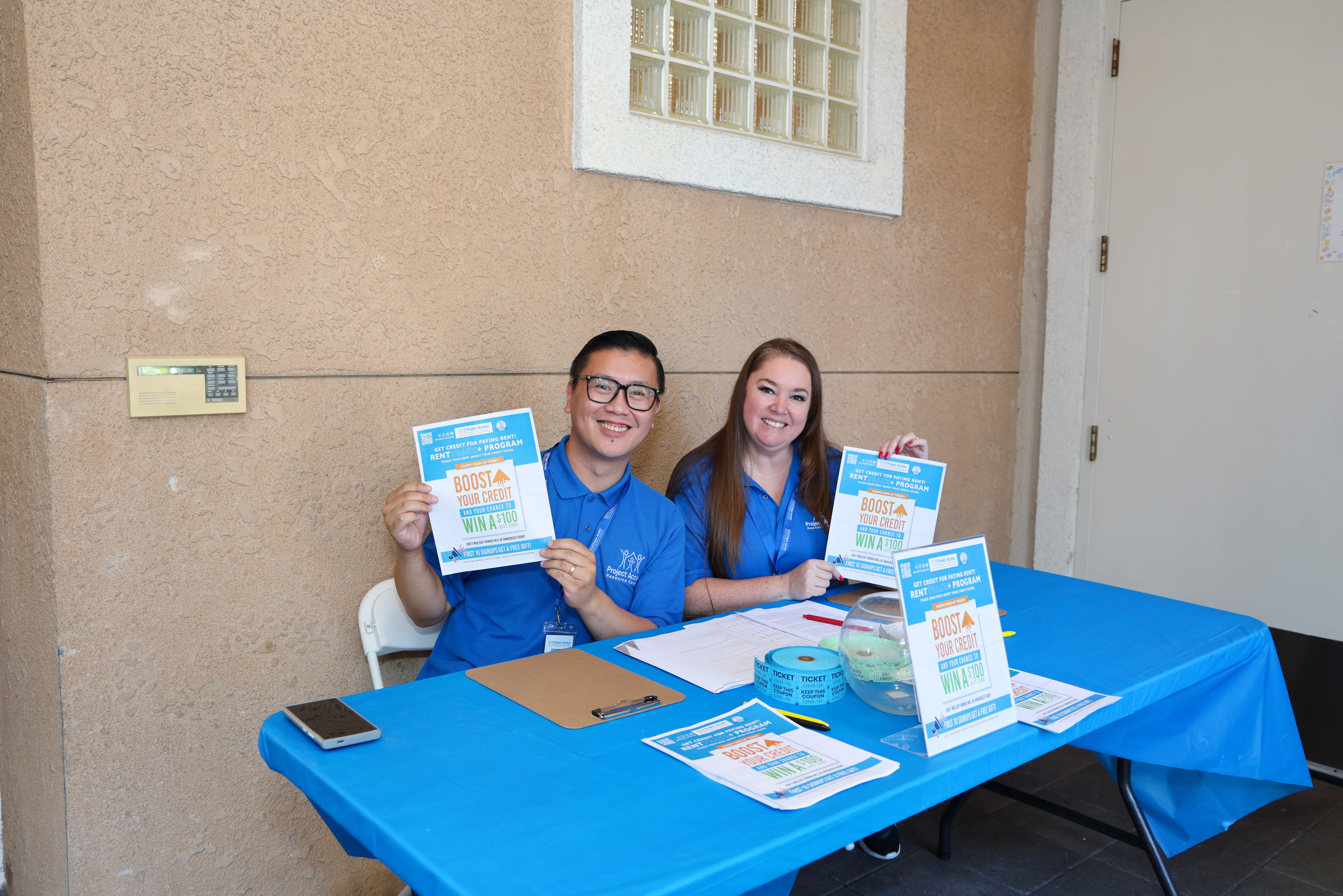 Children and adults play basketball together under a tent outside a building, while a man stands near the entrance watching.